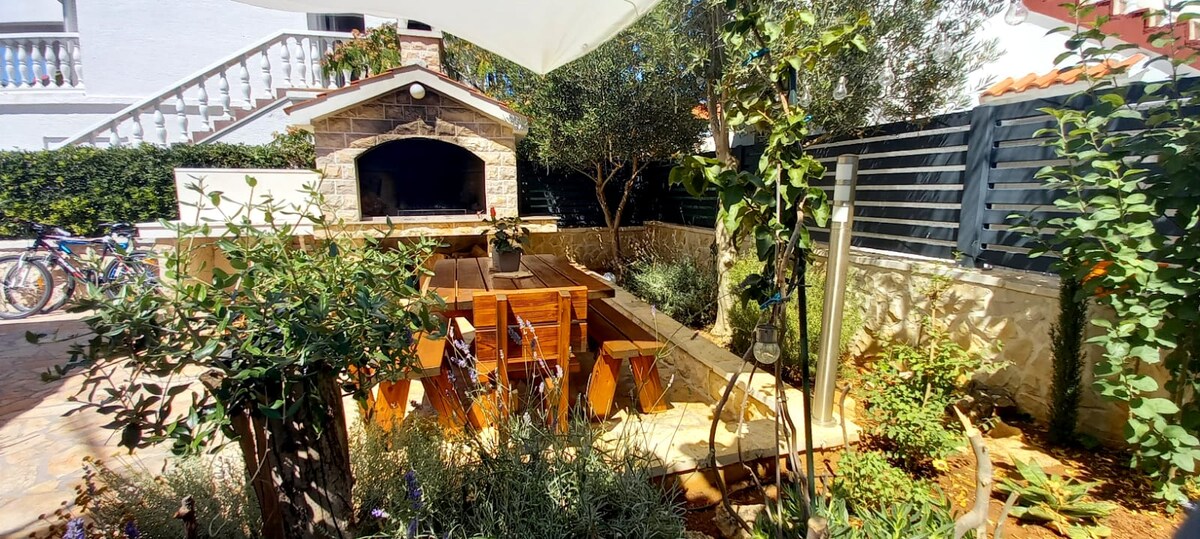 An outdoor dining area features a wooden table surrounded by chairs, set against a backdrop of greenery and floral accents. A stone grill is prominent, while the space is shaded by a white umbrella. Bicycles are visible in the background.