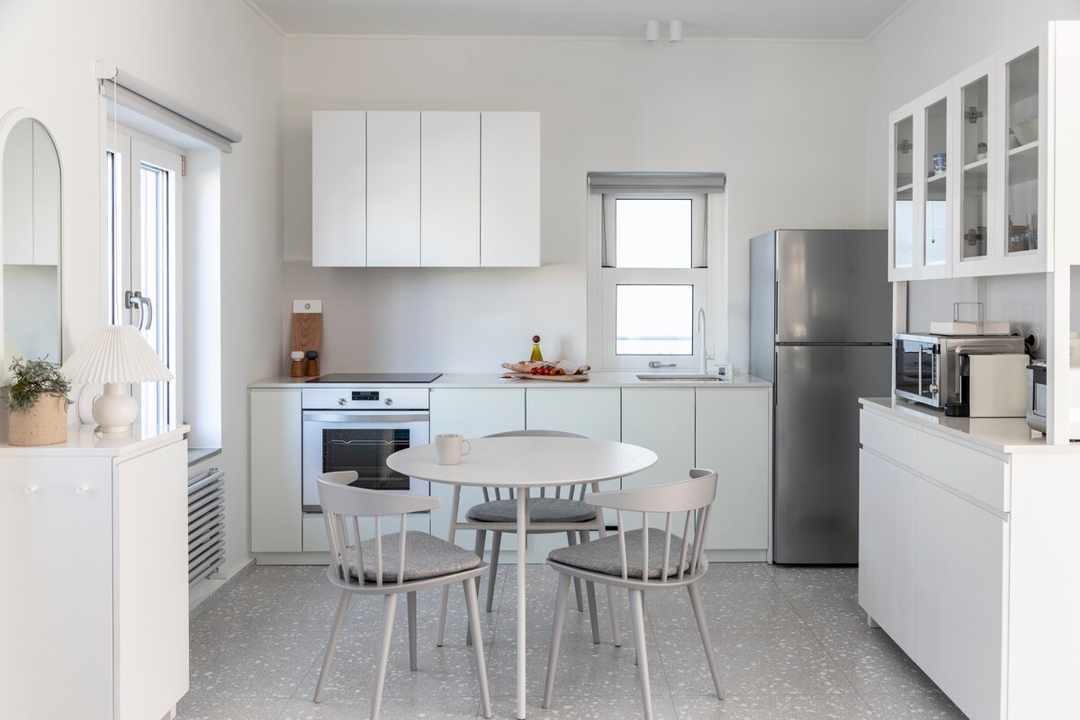 The kitchen features modern white cabinetry, a sleek countertop, and stainless steel appliances. A round dining table with four light grey chairs occupies the center. Natural light enters through two windows, enhancing the minimalist design of the space.