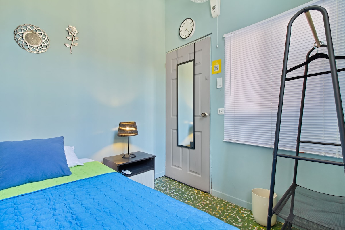 A simple room features light green walls and a bed with a blue coverlet, complemented by a blue pillow. A small lamp sits atop a nightstand next to the bed. The door leads to a corridor, and a folding chair is positioned nearby. A clock is mounted on the wall, and a window covered with blinds allows natural light to enter.
