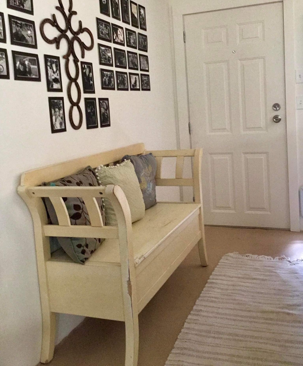 A light-colored wooden bench with an intricately carved backrest is positioned against a wall, adorned with a variety of framed black-and-white photographs. Decorative pillows in subtle patterns and colors are placed on the bench. A door with a circular handle is visible nearby, complementing the space.