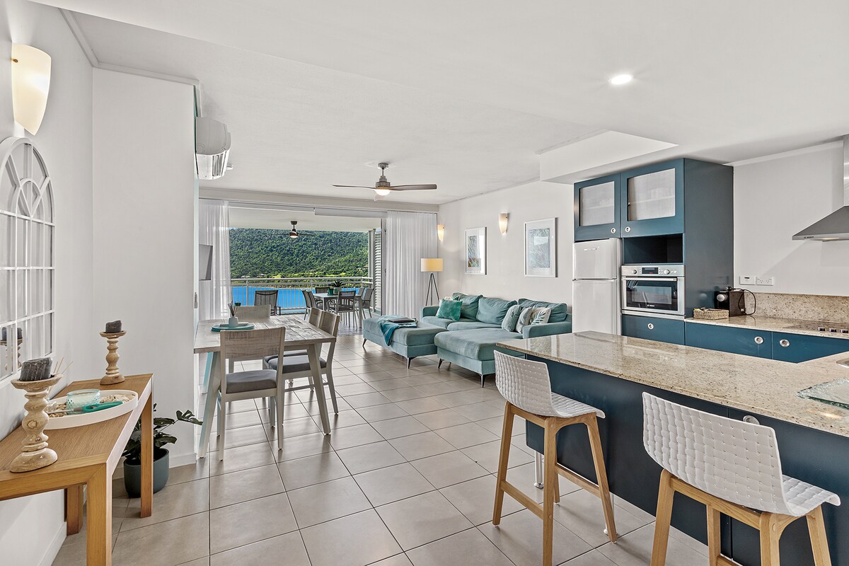 A spacious open-plan living area features a modern kitchen with granite countertops and built-in appliances. A dining table for six is positioned alongside light-colored seating, while large glass doors offer views of the landscape beyond, blending indoor comfort with the natural surroundings.