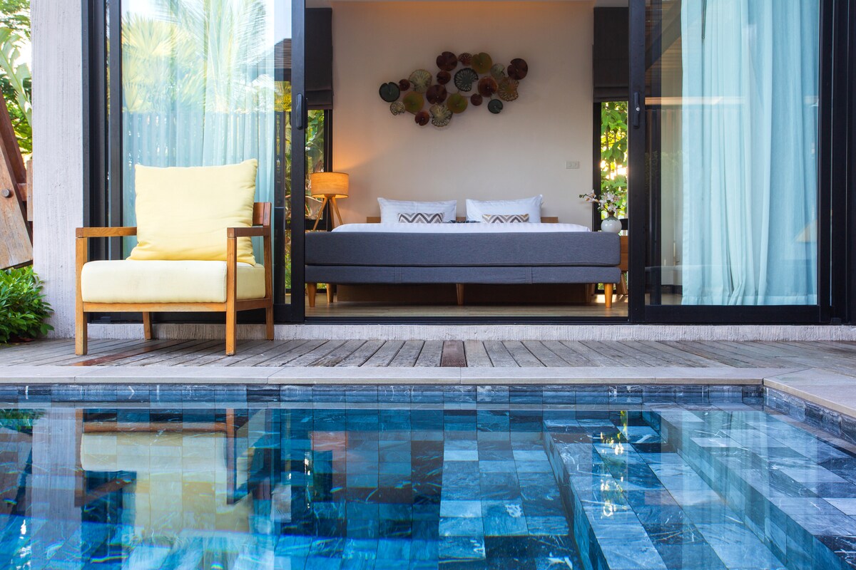 A private swimming pool reflects the clear sky, with a wooden deck surrounding it. Sunlight filters through the open sliding doors, revealing a well-decorated bedroom with a spacious bed and accent pieces. A yellow chair is positioned nearby, offering a relaxing spot with a view.
