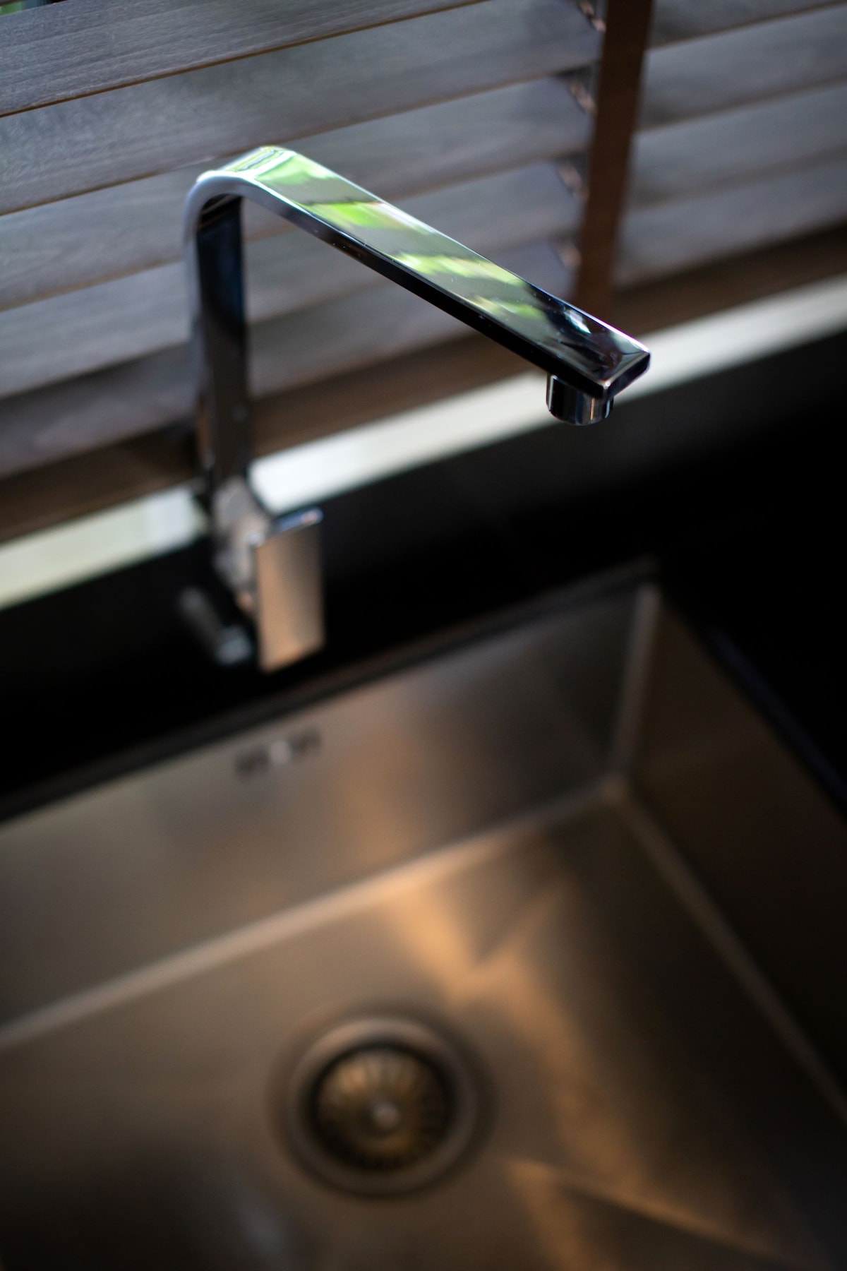 A modern kitchen faucet is positioned above a stainless steel sink, reflecting light from the surrounding area. The sink's polished surface complements the sleek faucet design, contributing to a contemporary kitchen aesthetic.