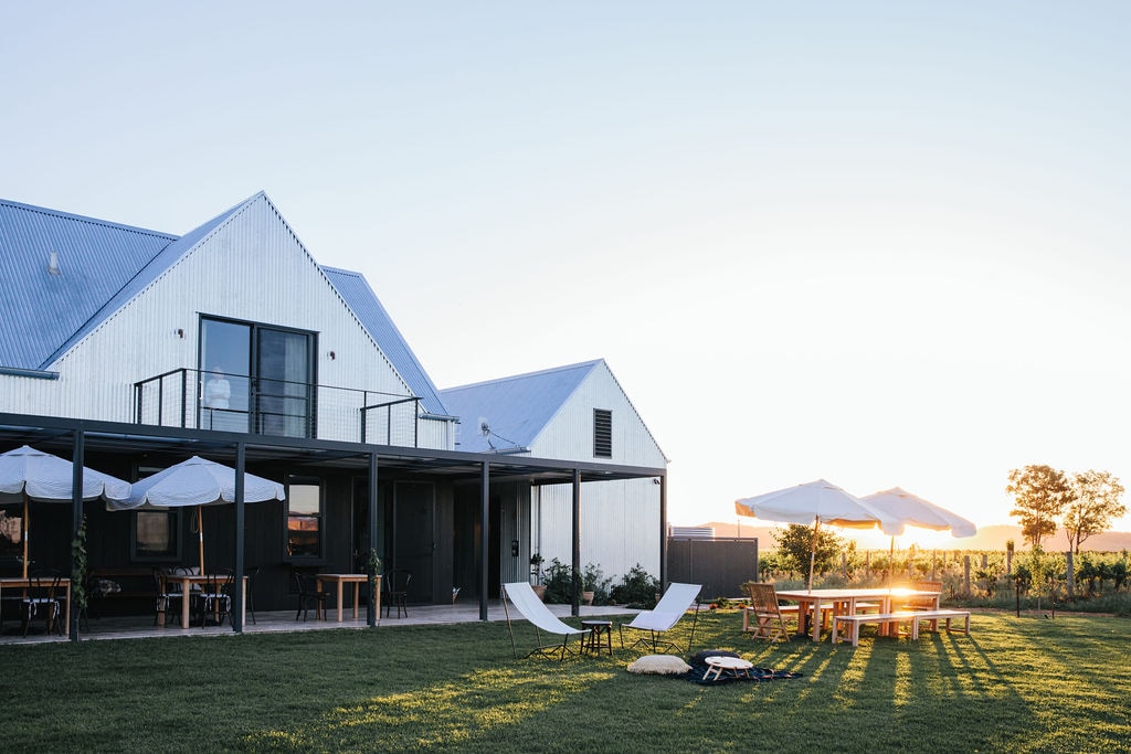 The outdoor area features a spacious lawn with sun loungers and a large dining table shaded by umbrellas. A striking sunset casts warm light over the modern building with a sleek metal exterior, showcasing large windows and a balcony that adds to the contemporary design.