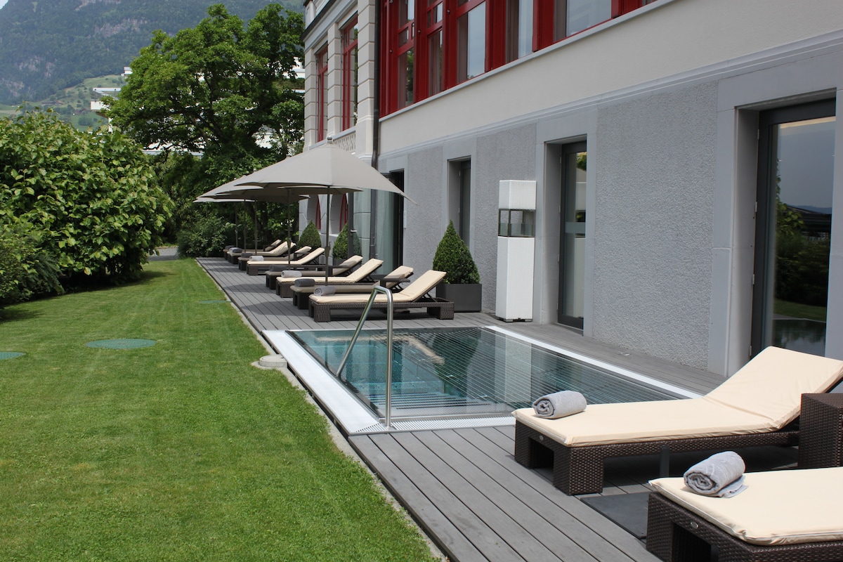 A sunlit terrace features a series of loungers arranged alongside a rectangular plunge pool. Shade is provided by large umbrellas, while a well-kept lawn and lush greenery create a serene environment. The building's modern architecture adds an elegant backdrop to this relaxation space.