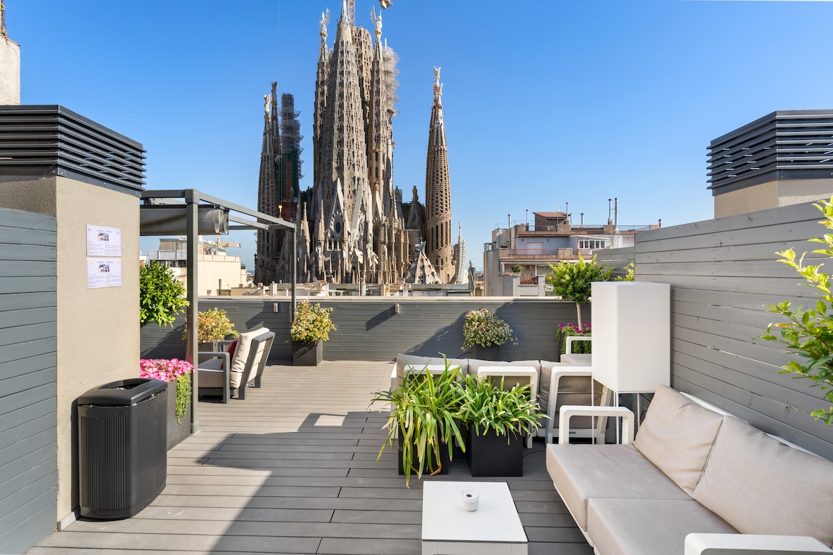 A spacious terrace presents views of the iconic Sagrada Familia, framed by green plants. Comfortable seating areas are arranged around a central table, providing a relaxing outdoor space. The terrace features modern design elements, with natural light enhancing the inviting atmosphere.