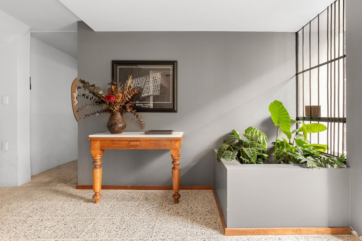 An entryway is featured with a wooden table and a framed artwork on a gray wall. A decorative vase with dried foliage is placed on the table. A green plant arrangement is situated in a built-in planter, providing a touch of nature to the space.