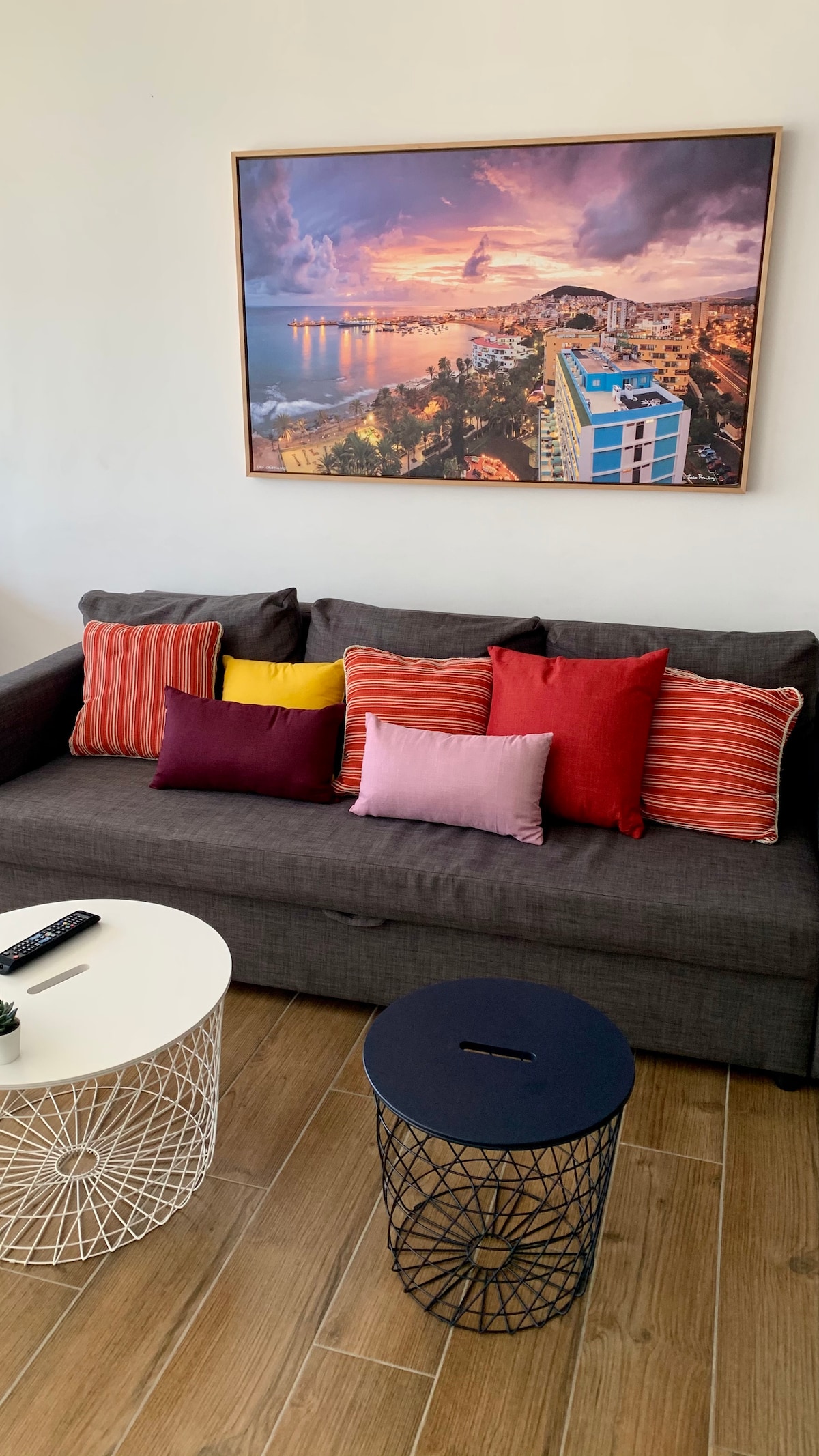 A lounge area features a comfortable sofa adorned with various colorful cushions in shades of red, yellow, and pink. A vibrant landscape photograph hangs above the sofa, capturing a scenic view. A white coffee table and a black wire stool complement the seating arrangement.