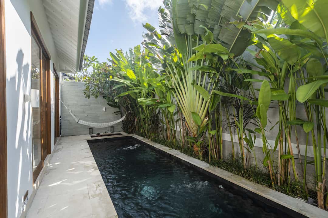 A serene private pool is surrounded by lush tropical greenery, featuring tall banana plants and carefully maintained foliage. The pool's dark water reflects the bright sky above, while white tiles create a clean border around the swimming area, enhancing the tranquil atmosphere.