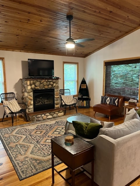 A welcoming living area is highlighted by a stone fireplace, featuring a mounted TV above it. Comfortable seating options include two chairs and a sofa, with a patterned area rug underfoot. Natural light filters through the windows, providing a bright and cozy atmosphere.