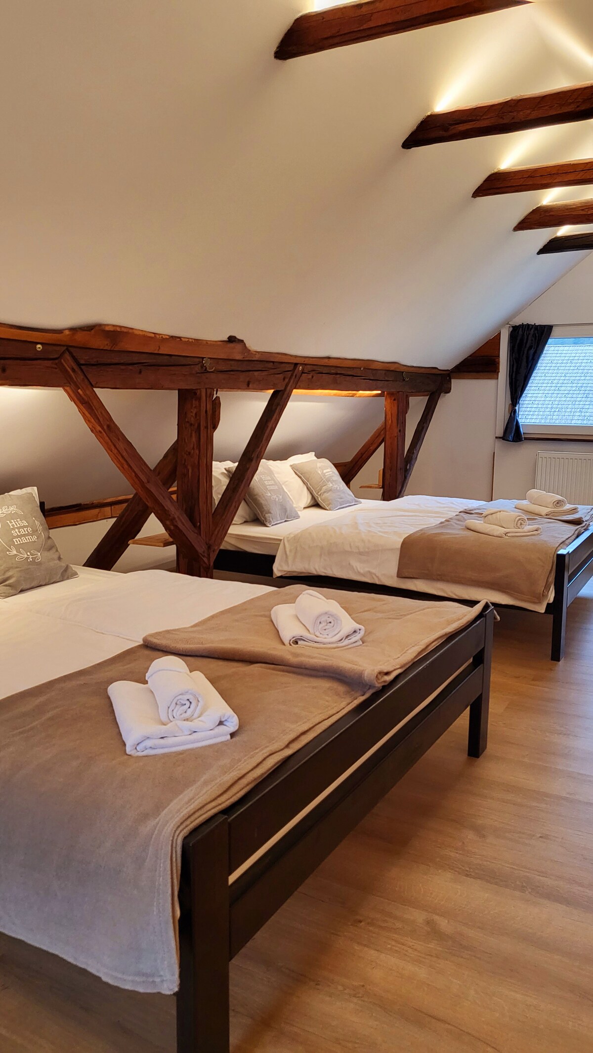 The spacious sleeping area is characterized by exposed wooden beams above and features two separate beds dressed in neutral linens. Soft towels are neatly rolled at the foot of each bed, and natural light filters through a window, highlighting the warm wooden tones of the floor.