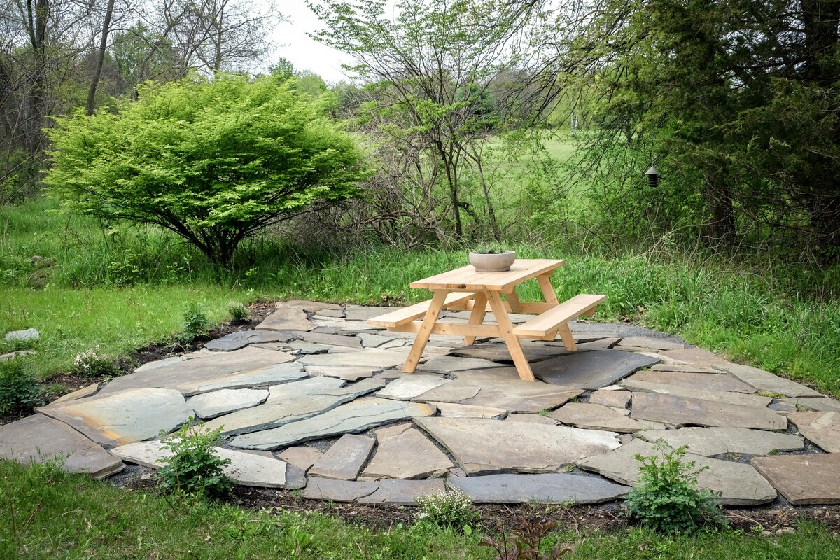 A stone patio is surrounded by lush greenery, featuring a simple wooden picnic table at its center. A low-growing shrub adds softness to the scene, while a grassy area extends beyond the patio, creating an inviting outdoor space for relaxation.