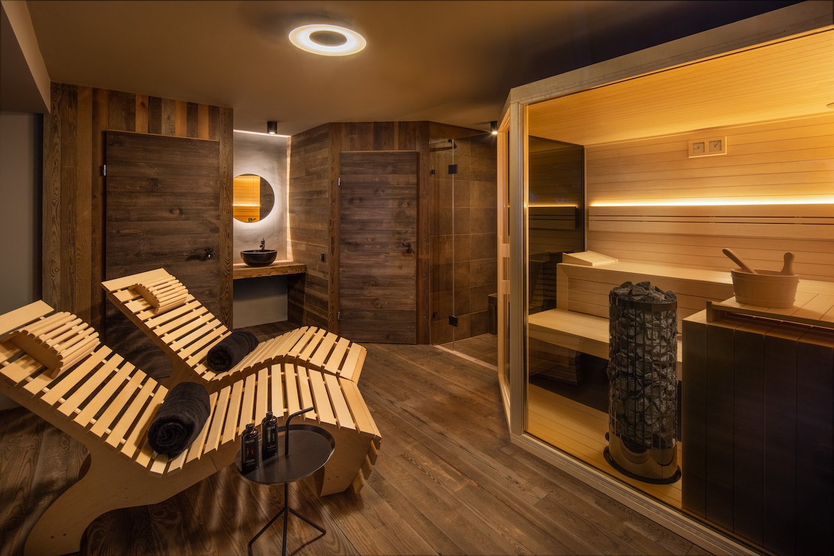 A soothing wellness area features two wooden loungers with rolled towels. The space is adorned with natural wood elements, illuminated by soft lighting. A sauna with a wooden interior is visible, complemented by a stone basin and circular mirror on the wall.