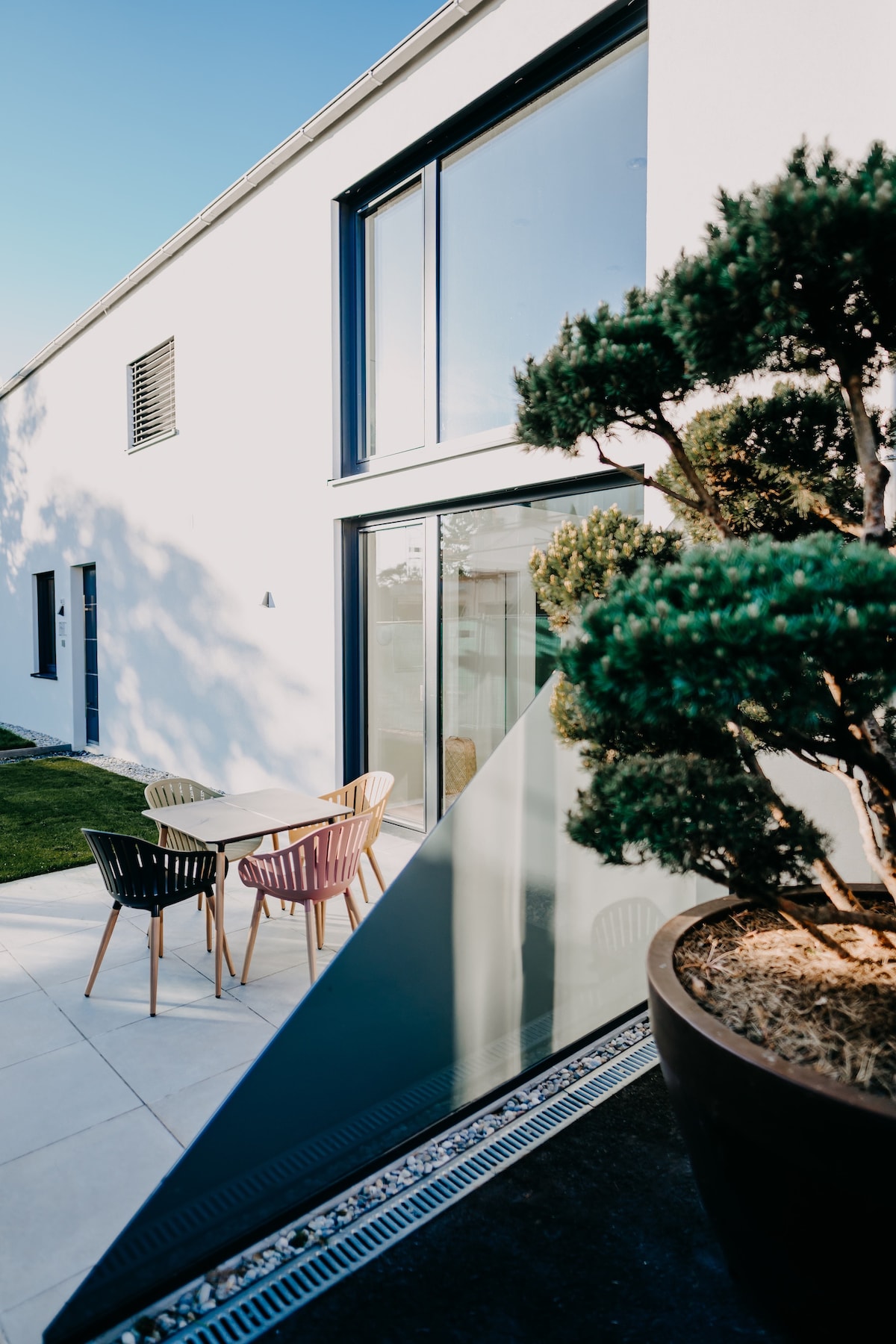 A modern exterior view of the chalet showcases a terrace with a small table and four chairs in varied colors. Lush greenery is present, including a sculptured tree in a large planter. Sunlight enhances the clean architectural lines and bright surface materials.