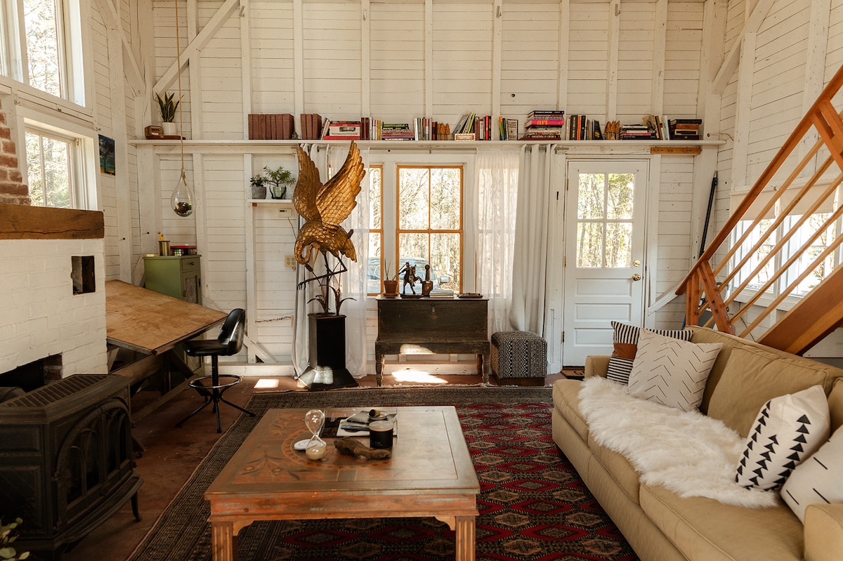 A cozy living area is highlighted by a spacious sofa adorned with a soft throw, accompanied by a wooden coffee table. Natural light streams through large windows, illuminating a rustic fireplace and a decorative sculpture. Bookshelves filled with various books create an inviting atmosphere.