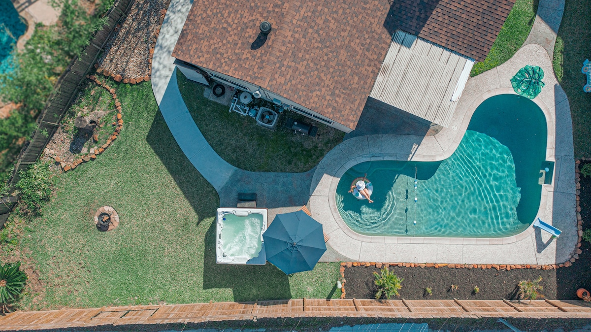 An aerial view captures a private pool with a curved design, surrounded by a spacious yard. A hot tub sits adjacent to the pool, accompanied by a shaded umbrella. The landscape is neatly maintained, with pathways leading to various outdoor areas.