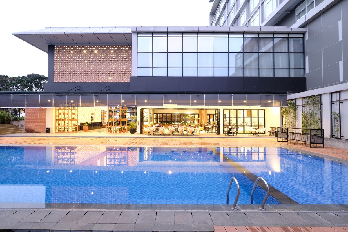 An outdoor swimming pool is framed by a modern hotel structure featuring large glass windows. The pool area reflects warm light from the restaurant inside, where both indoor and outdoor seating options are visible. Lush greenery is present along the pool's edge.