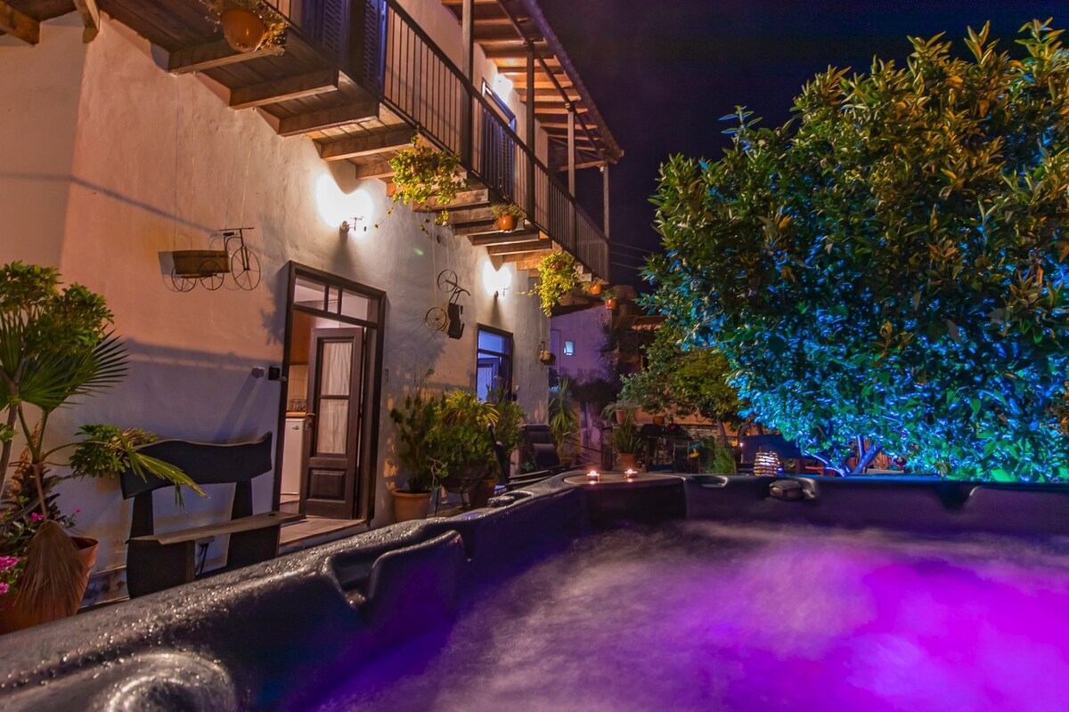 An outdoor hot tub is illuminated with soft purple lighting, surrounded by lush greenery and potted plants. A traditional building with wooden details and balcony is visible in the background, creating a tranquil ambiance ideal for relaxation.