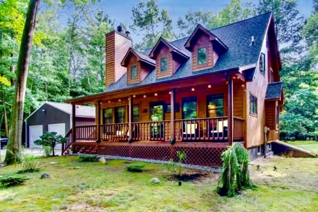 A charming wooden cabin features a spacious front porch with several seats, surrounded by lush greenery. The exterior showcases a combination of dark wood and paneling, with multiple gabled roof sections and large windows that invite natural light.