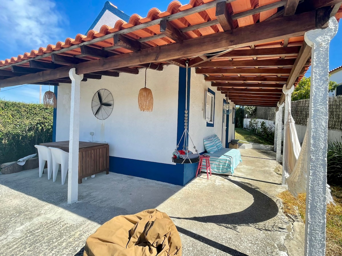A covered outdoor area features a wooden table with four white chairs, alongside a hammock and a lounge chair. The roof is supported by wooden beams, providing shade. A light fixture hangs from above, and a small red stool adds a pop of color.