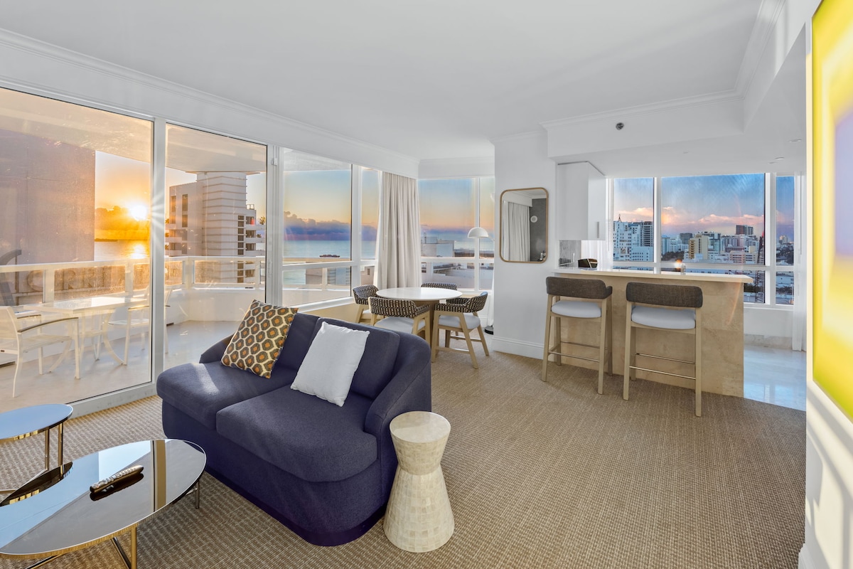 A modern living area is highlighted by large glass windows that capture a vibrant sunset view. Comfortable seating and a sleek table are arranged, with a dining area and kitchen bar visible in the background. Neutral tones create an open and airy atmosphere.