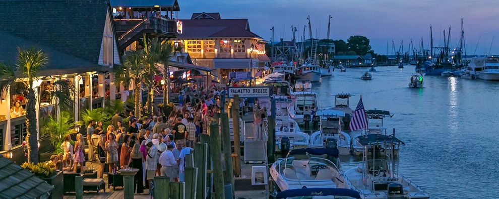 A vibrant waterfront scene is depicted, featuring numerous boats lined along a lively dock. Stalls and restaurants are bathed in warm light, with groups of people enjoying the outdoor space. The background showcases a variety of boats and masts, all under a dusky, colorful sky.