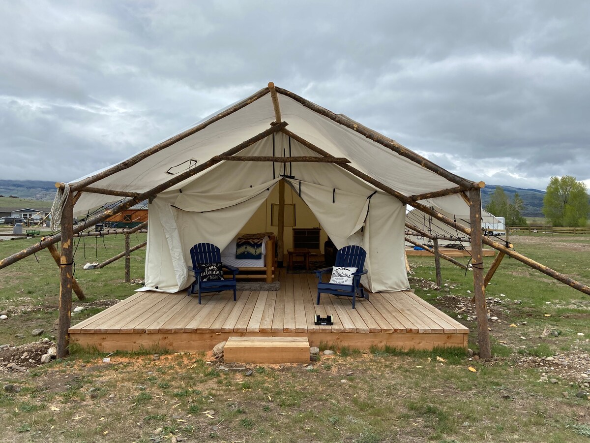 A spacious glamping tent is set on a wooden deck, featuring two blue adirondack chairs positioned in front. The canvas exterior is supported by wooden poles, while interior furnishings are visible through the entrance, creating a welcoming outdoor space.