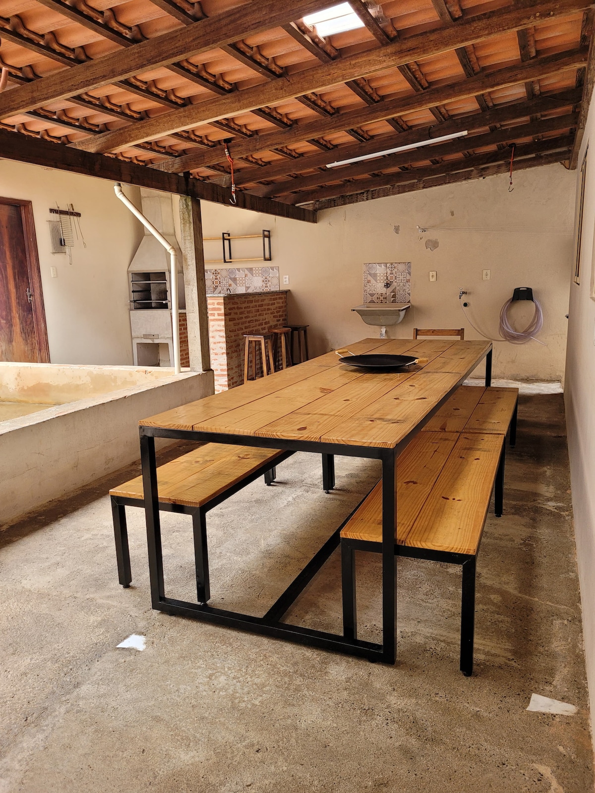 An outdoor dining area features a long wooden table surrounded by benches. A simple light fixture is suspended from the ceiling, illuminating the space. Walls are unfinished, and a small sink is visible in the corner, contributing to the casual ambiance.