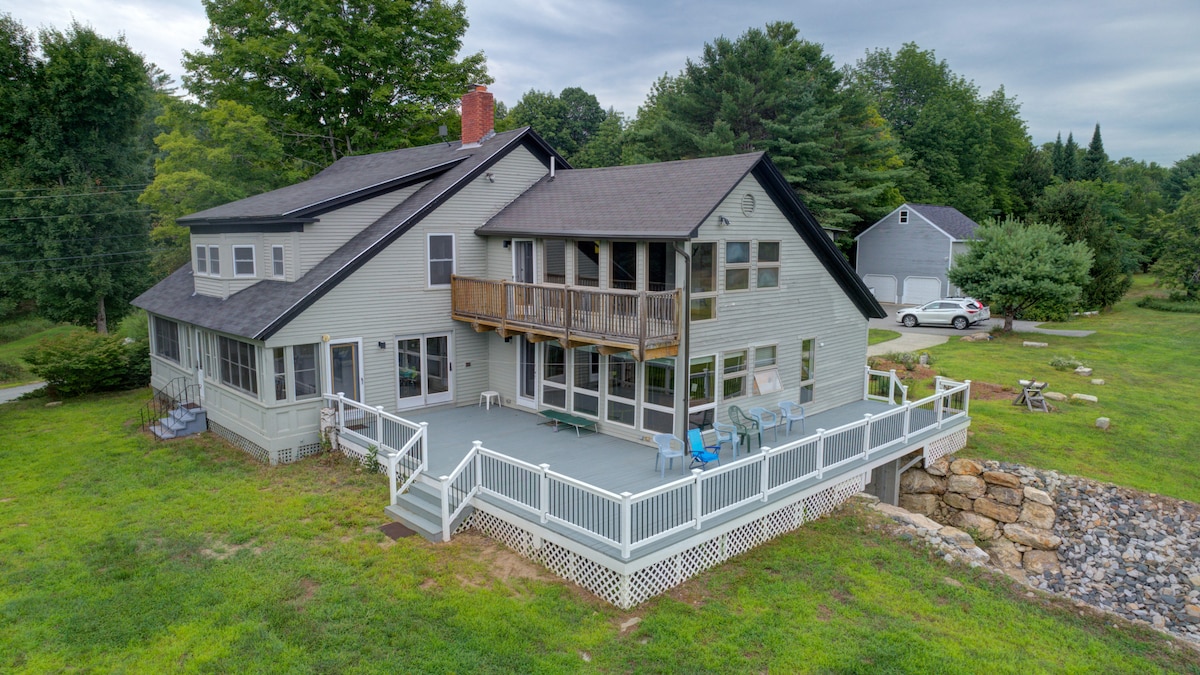 The farmhouse is set amidst a lush green landscape, featuring a spacious deck that wraps around part of the building. Large windows allow natural light in, while a blend of outdoor seating areas invites relaxation. The property is bordered by trees and open spaces, creating a serene environment.