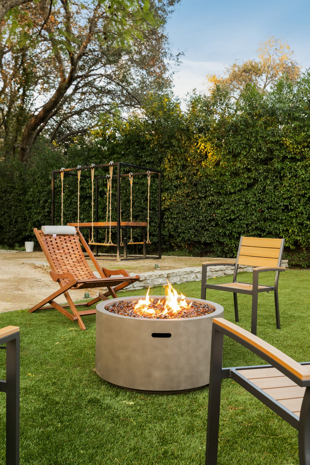 An outdoor patio area features a circular fire pit surrounded by four chairs. The settings include two wooden reclining chairs and two metal-framed chairs with wooden slats. A grassy area is present, along with a swing set in the background, providing a relaxing atmosphere.