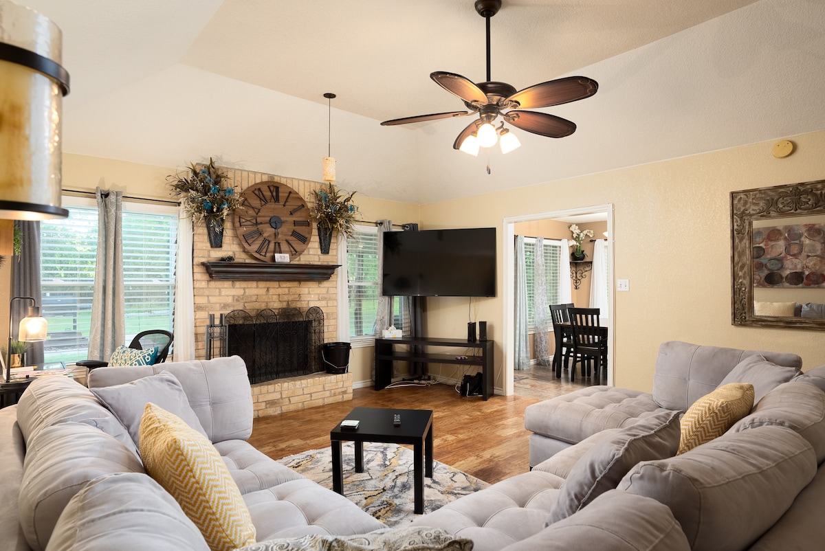 A spacious living area features a large, plush sectional sofa with accent pillows facing a flat-screen television. The exposed brick fireplace serves as a focal point, while large windows provide natural light and views of the outdoors. Decorative elements include a wall clock and plants.