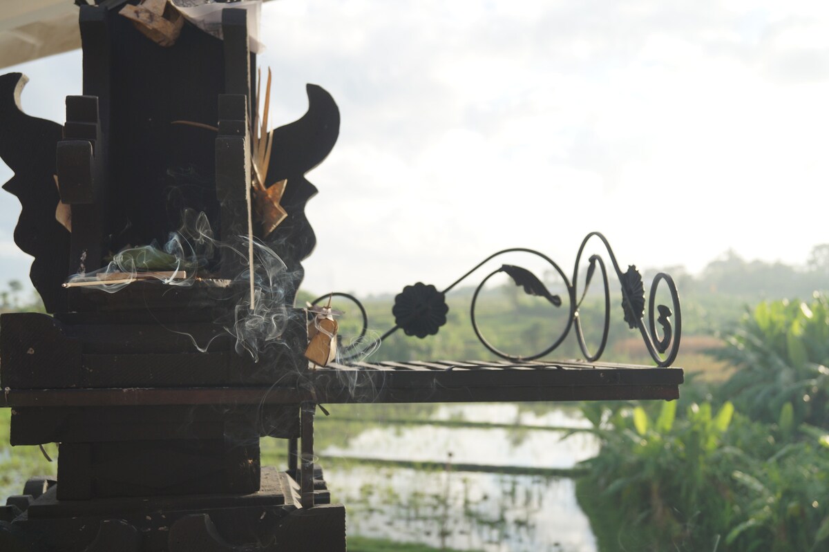 A traditional Balinese offering stand is depicted with curling smoke rising from an offering, set against the backdrop of a rice field. Lush greenery is visible in the distance, while soft, natural light illuminates the scene.