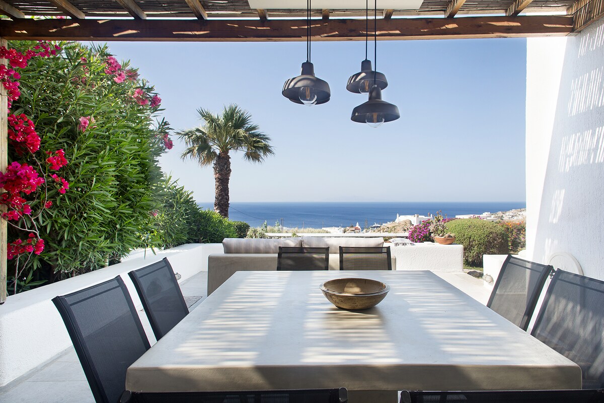 An outdoor dining area presents a spacious table surrounded by black mesh chairs, beneath wooden slats providing partial shade. The view features lush greenery with bright flowers and a scenic backdrop of the Aegean Sea, enhancing the tranquil setting.