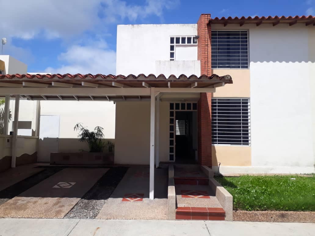The image displays a two-story building with a shaded entrance. A tiled walkway leads to the front door, framed by a small green lawn and a low plant. Large windows are positioned on either side, allowing natural light into the space.