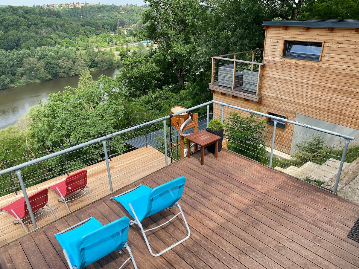 An outdoor deck features two blue lounge chairs and a small wooden table, providing a space for relaxation. The surrounding greenery and the view of the river below enhance the peaceful atmosphere. A wooden structure is visible in the background, adding to the natural setting.