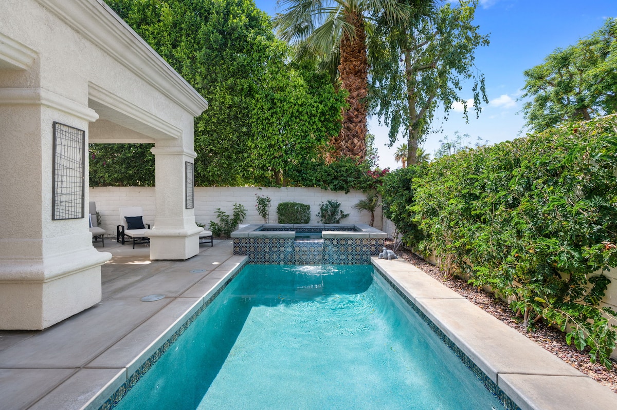 A private swimming pool is surrounded by lush greenery, featuring a spa at one end. Sunlight reflects off the clear water, while comfortable lounge chairs are placed nearby, inviting relaxation. A covered patio area can also be seen, enhancing the outdoor space.