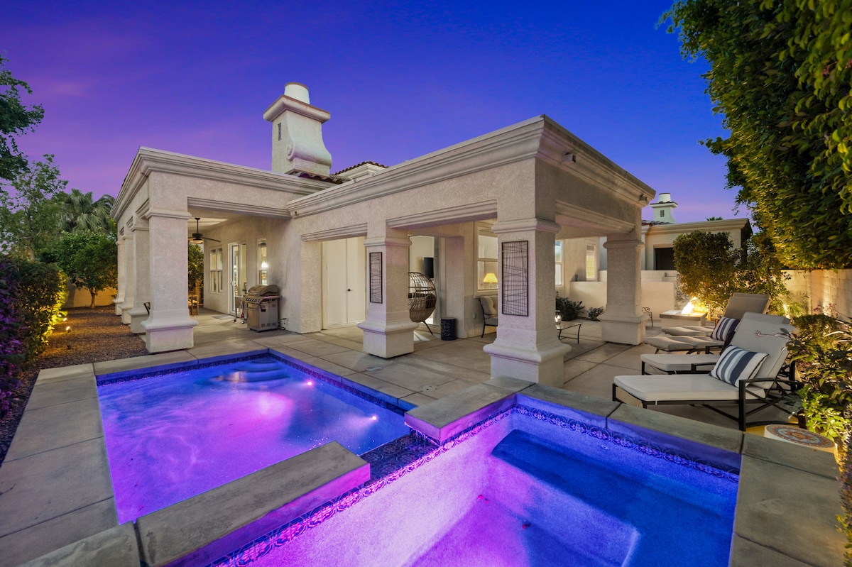 An outdoor space features a private pool and hot tub, illuminated in soft purple hues, surrounded by elegant lounge chairs. The home is showcased with architectural details and partially shaded areas under a warm evening sky.