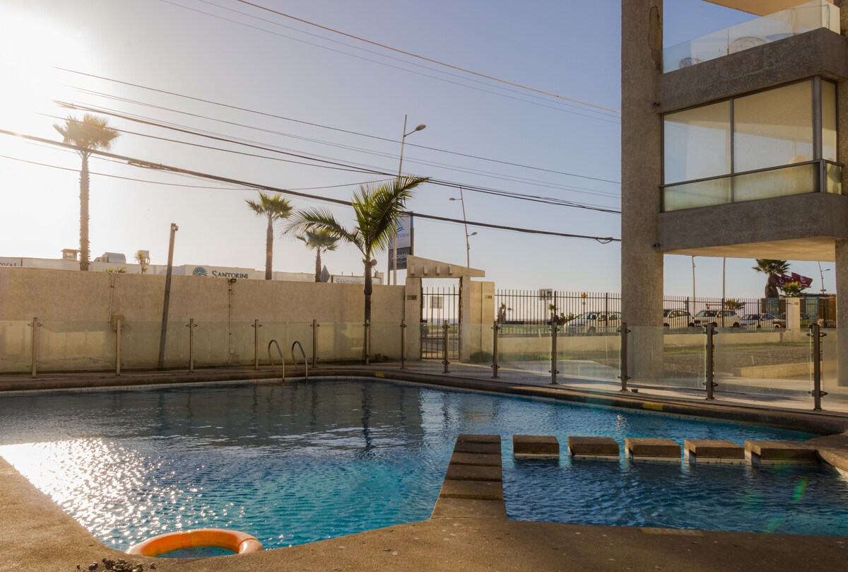 A swimming pool is positioned at the forefront, reflecting sunlight under a clear sky. Palm trees are visible in the background, offering a sense of space and relaxation. The surrounding area is fenced, ensuring privacy, while access points to the pool are indicated by steps.