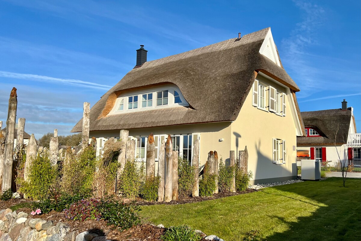 The villa features a traditional thatched roof and a light-colored exterior. A lush grassy area surrounds the home, with natural stone elements and carefully manicured flowerbeds. Clear skies are visible above, enhancing the inviting appearance of the property.