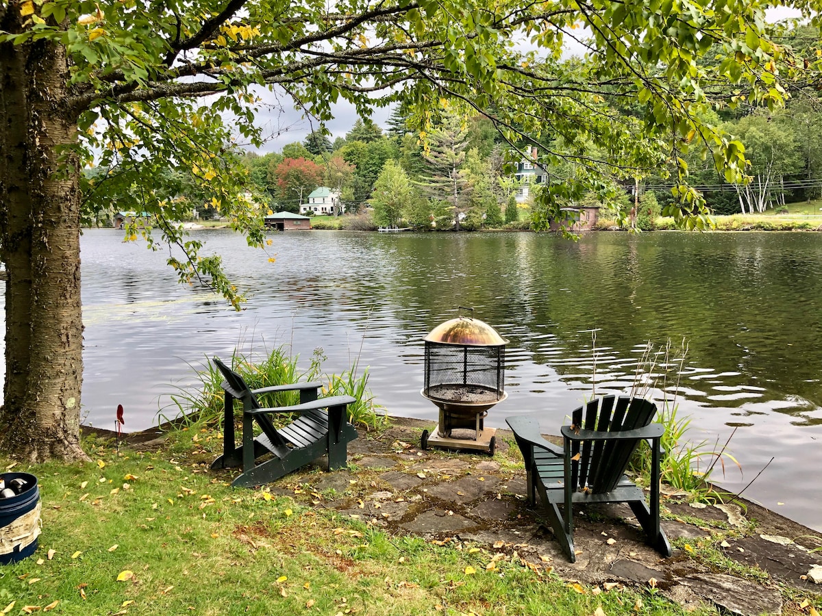 Two Adirondack chairs are positioned near the lakefront, accompanied by a fire pit on a stone patio. Lush greenery frames the scene, with gentle water ripples reflecting the surroundings. A peaceful view includes the lake and distant houses beyond the water.
