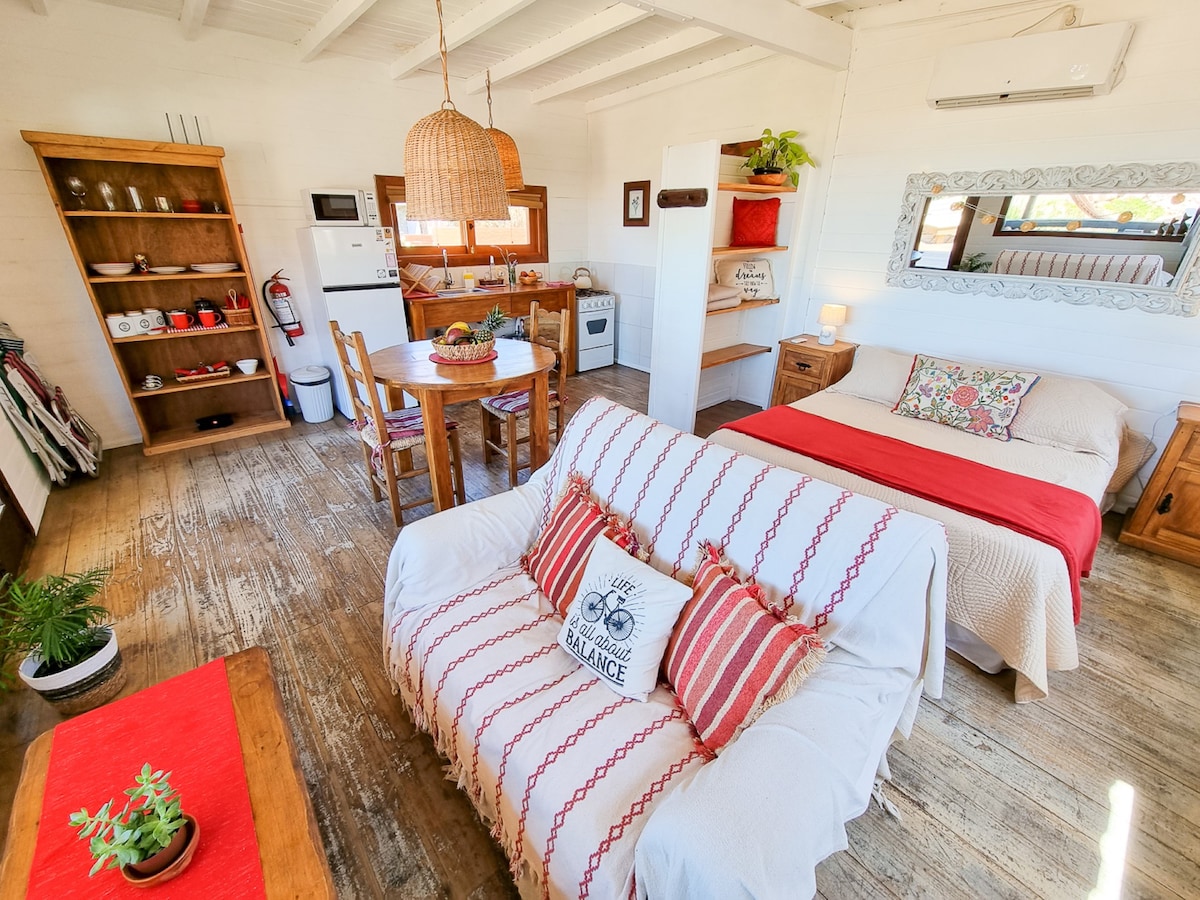 The open-plan living area features a spacious bed covered with neutral bedding, complemented by decorative cushions. A comfortable sofa is adorned with a patterned throw. A dining table with chairs is positioned nearby, and wooden shelves display kitchen essentials. Natural light fills the space from windows.