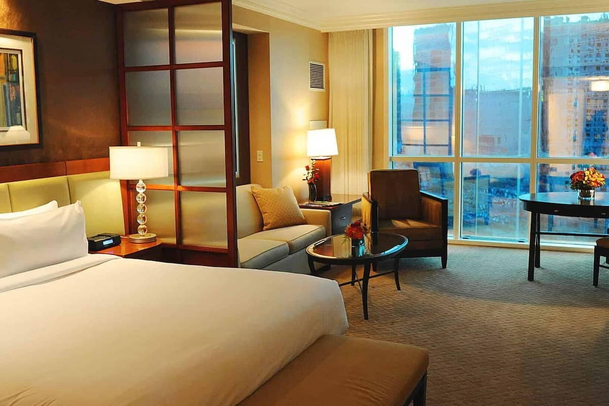 A spacious studio suite features modern furnishings, including a king bed with crisp white linens. A comfortable seating area is set up with a sofa and two chairs, while large windows provide natural light and views of the surroundings. A round table adds a cozy touch.