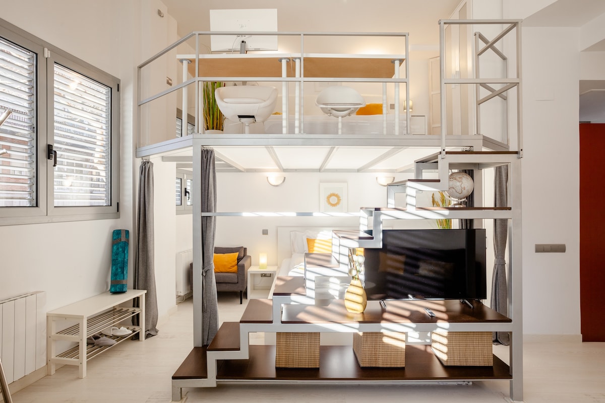 A modern loft space features a raised platform with two sinks above and a comfortable seating area below, illuminated by natural light from the windows. Open shelving reveals decorative accents, while a television is positioned on a sleek media console.