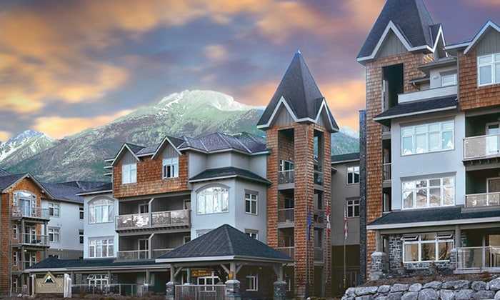 The exterior of the building features a blend of modern and traditional architecture. Natural stone and wood accents are visible, complemented by sloped roofs. The backdrop includes majestic mountains under a colorful sky, enhancing the overall scenic appeal.