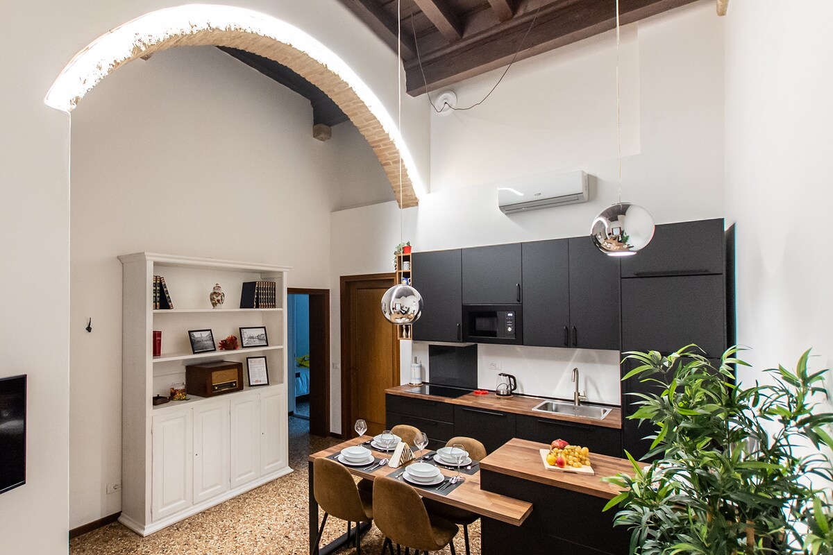 A modern kitchen area is presented, featuring black cabinetry and a wooden dining table set with dishes and fruit. A stylish arch highlights the ceiling, while decorative elements and natural greenery create a balanced atmosphere. Stainless steel appliances are visible for convenience.