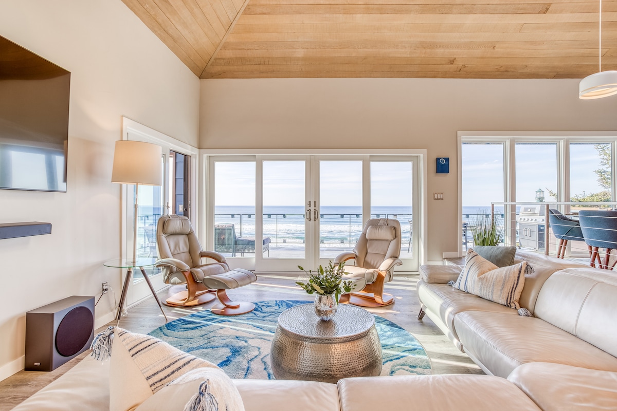 A spacious living area features large glass doors that frame scenic ocean views. A light-colored sectional sofa surrounds a round metallic coffee table, while a textured area rug adds warmth. Natural light floods the space, accentuating the high wooden ceiling and coastal decor.