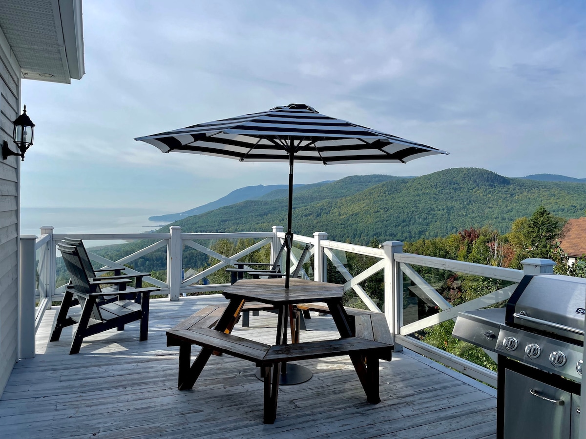 A spacious outdoor deck is equipped with a large striped umbrella, providing shade over a wooden picnic table and adjacent seating. A barbecue grill is positioned nearby, with sweeping views of the lush mountains and approachable waters of the St. Lawrence River in the background.