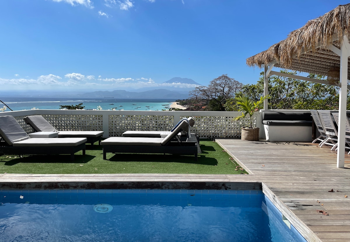 A wooden deck surrounds a clear blue pool, with lounge chairs positioned alongside. In the background, views of the coastline and distant mountains are framed by a clear blue sky. A shaded seating area is visible to one side, enhancing the outdoor space.
