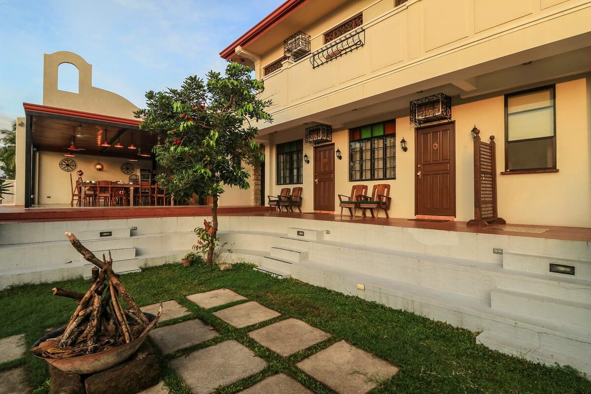 A spacious outdoor area features a well-maintained garden, with stone pathways leading to an entrance flanked by wooden chairs. A pergola seating area is visible in the background, offering an inviting space for gatherings. A fire pit with stacked firewood is also present.