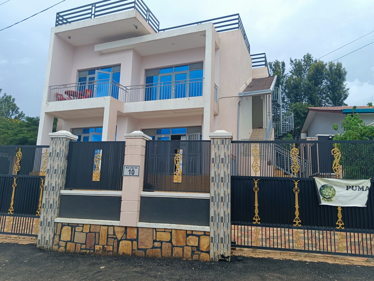 The exterior of Puma Garden B&B is showcased, featuring a two-story structure with large glass windows and a spacious balcony. A decorative gate, fashioned from wrought iron, surrounds the property, while stonework lines the base of the building, providing a solid foundation.